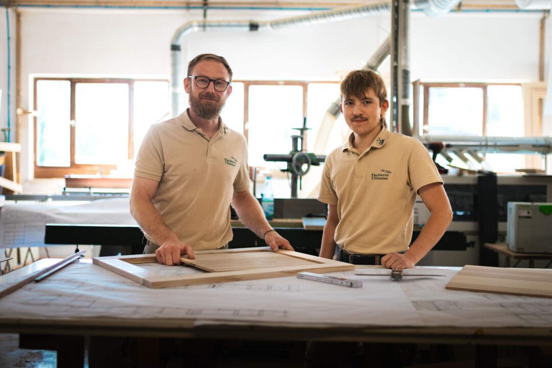 Zwei Tischler stehen in ihrer Werkstatt hinter einem Arbeitstisch. Sie blicken beide in die Kamera, während der ältere Mann einen unfertigen Holzrahmen hält.