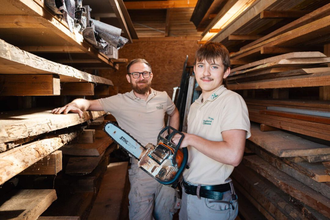Zwei Tischler in ihrer Werkstatt. Der ältere Mann steht lächelnd neben einer Holzpalette, während der jüngere eine Kettensäge hält und in die Kamera blickt.