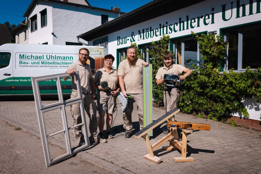 Das Team der Tischlerei Uhlmann, in Summe vier Personen, posiert lächelnd vor ihrer Werkstatt. Sie halten verschiedene Werkzeuge und ein Fensterrahmen.