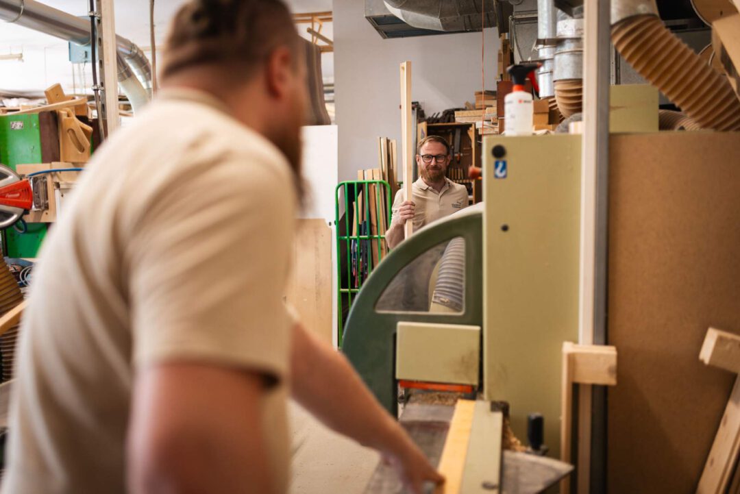 Ein lächelnder Tischler schaut aus einem großen Holzbearbeitungsgerät hervor. Ein unscharfer Kollege im Vordergrund hält ein Holzstück.