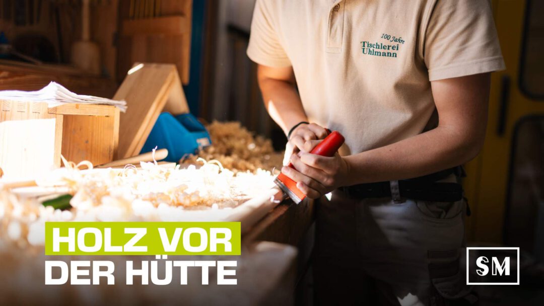 Ein Tischler bei der Arbeit, wie er einen Meißel verwendet, um Holzspäne zu entfernen. Der Fokus liegt auf seinen Händen und dem Werkzeug. Im Vordergrund steht der Text "HOLZ VOR DER HÜTTE".