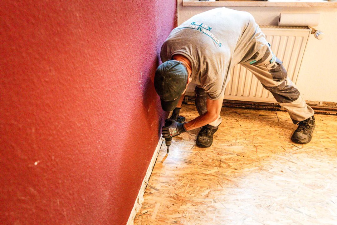 Handwerker (SM Kattner) mit Baseballcap verschraubt eine OSB-Platte am Rand des Fußbodens an einer roten Wand, um den stabilen Fehlboden fertigzustellen und das Ausrieseln der Schüttung zu verhindern.