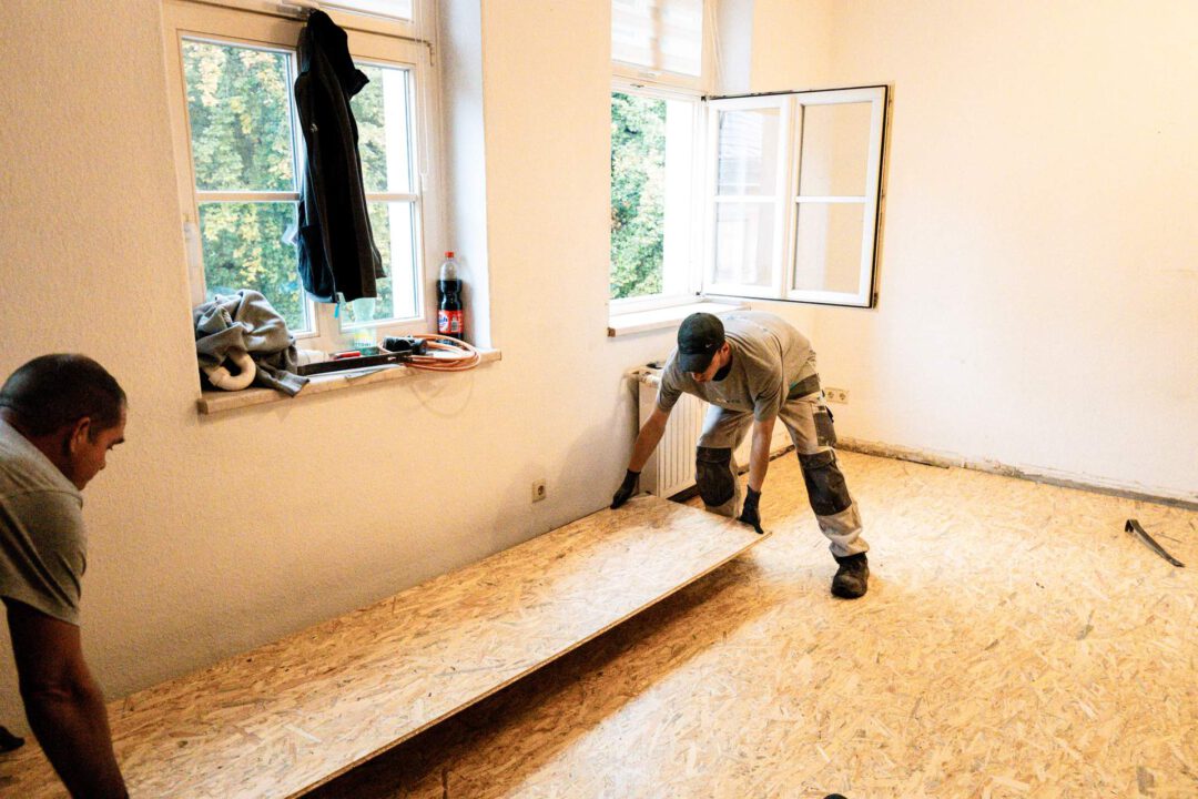 Zwei Handwerker (SM Kattner) bei der Bodenverlegung von OSB-Platten auf einer Baustelle in Lößnitz. Die Platte wird als Fehlboden im Zuge einer Beseitigung eines Wasserschadens verlegt.