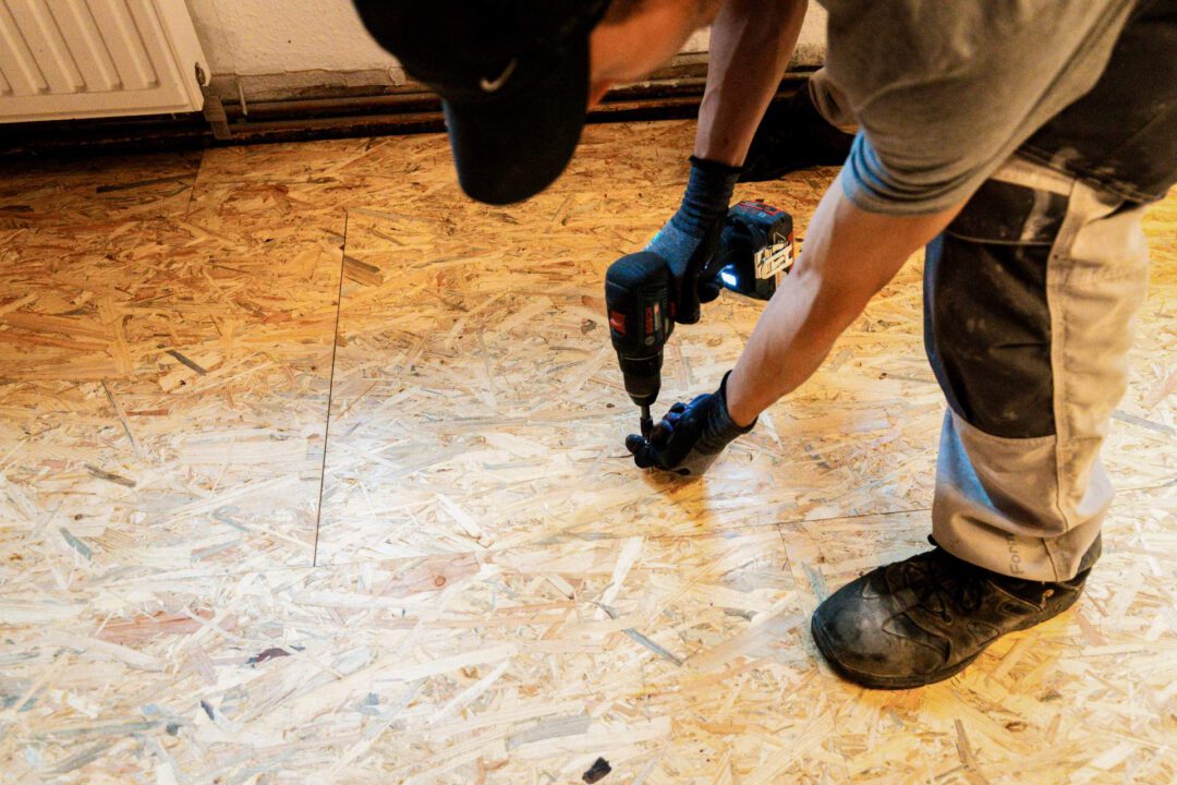Handwerker der Firma SM Kattner befestigt eine OSB-Platte mit einem Akkuschrauber auf dem neu eingebrachten Fehlboden zur weiteren Stabilisierung des Untergrunds für den neuen Laminatboden.