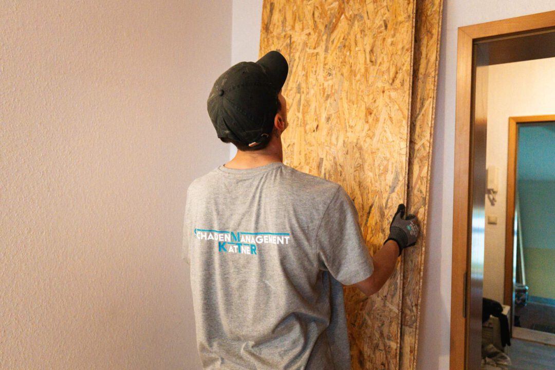 Handwerker (Rückenansicht) in einem T-Shirt der Firma Schadensmanagement Kattner (SM Kattner) hält eine OSB-Platte an eine Wand. Die Platte wird möglicherweise als Wandverkleidung oder zur Stabilisierung verwendet.