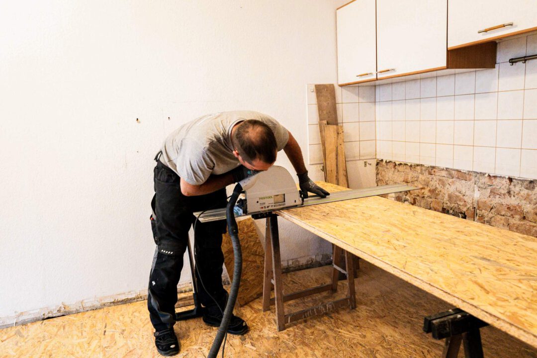 Handwerker der Firma SM Kattner beim Zuschnitt einer OSB-Platte mit einer Tauchsäge in einer im Umbau befindlichen Küche. Die Platten dienen zur Begradigung und Stabilisierung des Bodens nach dem Einbringen der Schüttung.
