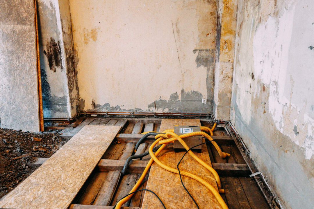 Ausschnitt des Bodens während der Sanierungsarbeiten: Freiliegende Holzbalken der Unterkonstruktion und Teile des Bodens, bedeckt mit OSB-Platten. Gelbe Schutzschläuche liegen im Zentrum.