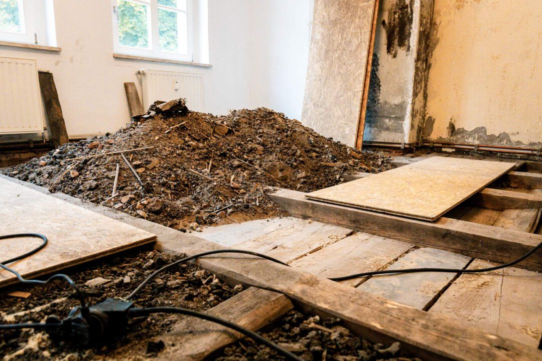 Baustellenansicht in Lößnitz nach einem Wasserschaden. In der Mitte des Raumes liegt ein großer Haufen der entfernten alten Bodenschüttung auf den freigelegten Holzbalken. Rechts und links sind bereits neue OSB-Platten zur Herstellung des stabilen Fehlbodens durch SM Kattner platziert.