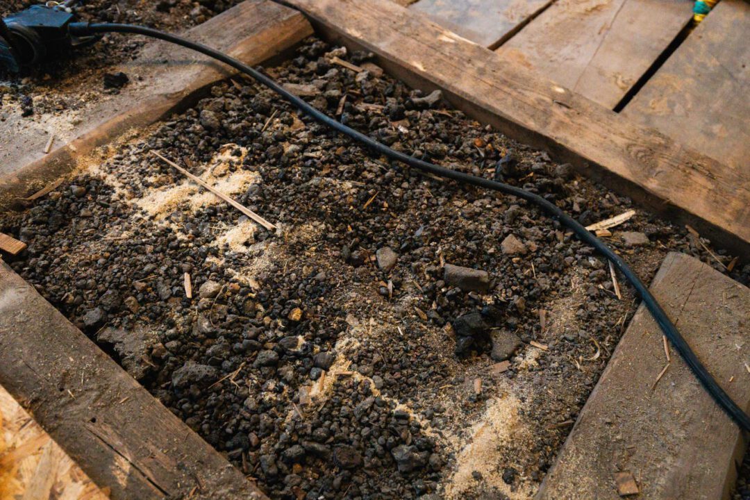 Nahaufnahme der Bodenschüttung (möglicherweise Lavasplit oder mineralische Schüttung) zwischen den Holzbalken der Unterkonstruktion. Diese Schüttung wird bei der Bodensanierung durch SM Kattner zur Isolierung und Begradigung des Fehlbodens vor der Verlegung der OSB-Platten in Lößnitz eingesetzt.