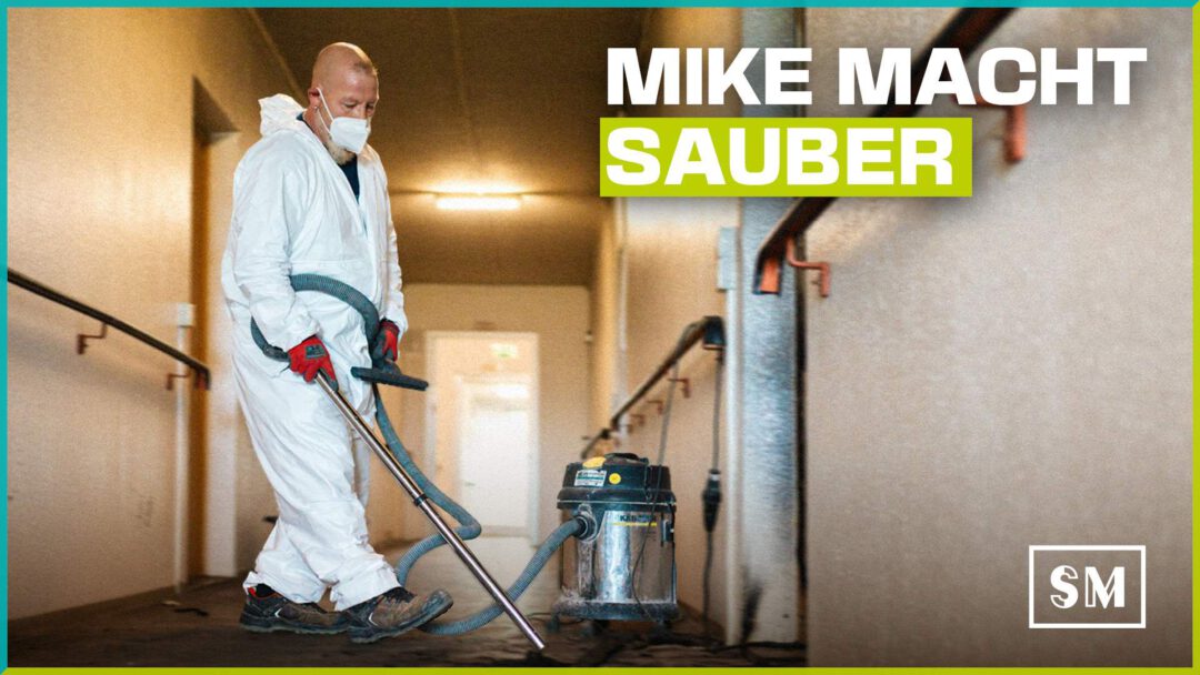 Social-Media-Grafik: Ein Arbeiter namens Mike in weißem Schutzanzug und Atemschutzmaske saugt Brandschutt in einem Flur mit einem Industriesauger. Text-Overlay: "MIKE MACHT SAUBER"
