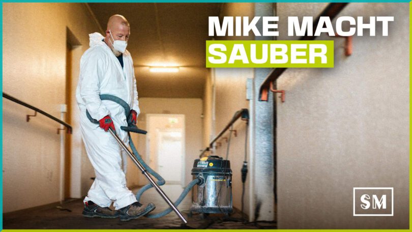 Social-Media-Grafik: Ein Arbeiter namens Mike in weißem Schutzanzug und Atemschutzmaske saugt Brandschutt in einem Flur mit einem Industriesauger. Text-Overlay: "MIKE MACHT SAUBER"