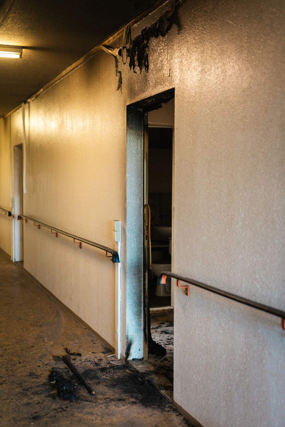 Seitenansicht eines verrußten Flurs mit Handläufen an den Wänden. Im Bildvordergrund ist ein durch Brand beschädigter Türrahmen zu sehen, der ins Innere eines Zimmers führt. Der Boden ist mit Ruß bedeckt.