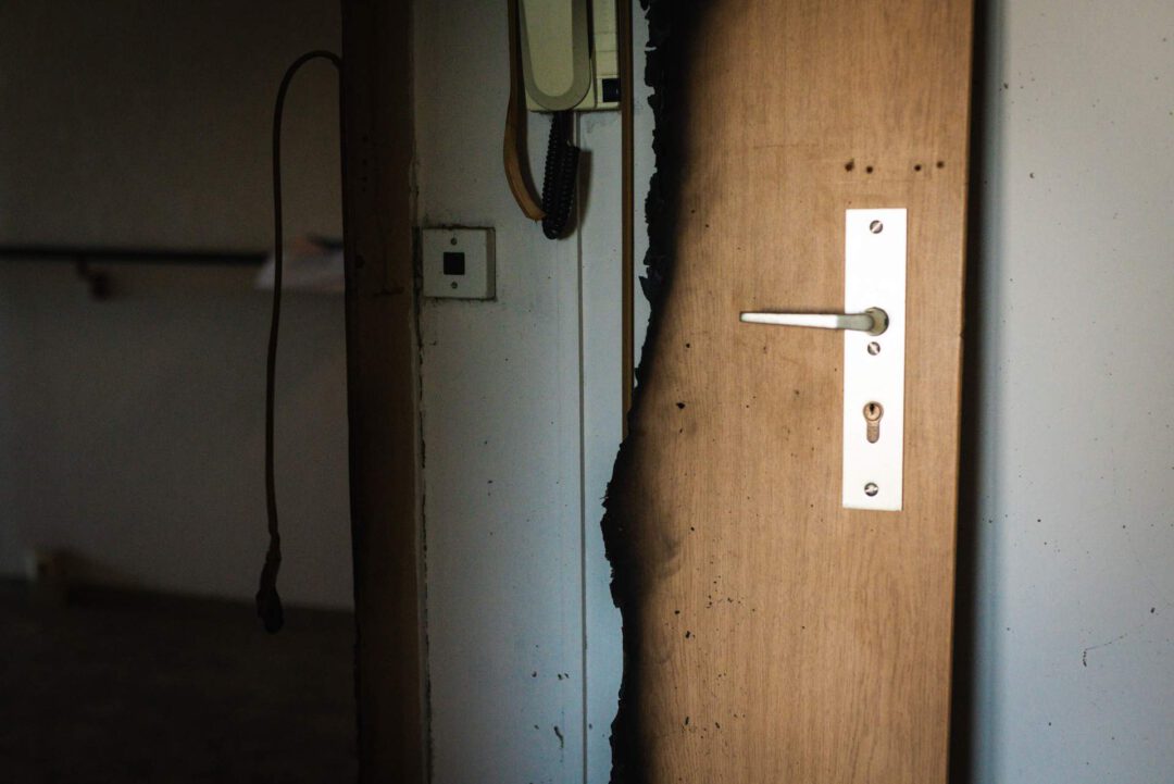 Nahaufnahme einer durch einen Brand beschädigten Holztür und Türrahmen mit deutlichen Rußspuren und einem verbrannten Rand. Man sieht ein Türschloss, einen Türgriff und eine Gegensprechanlage/Steckdose an der Wand.