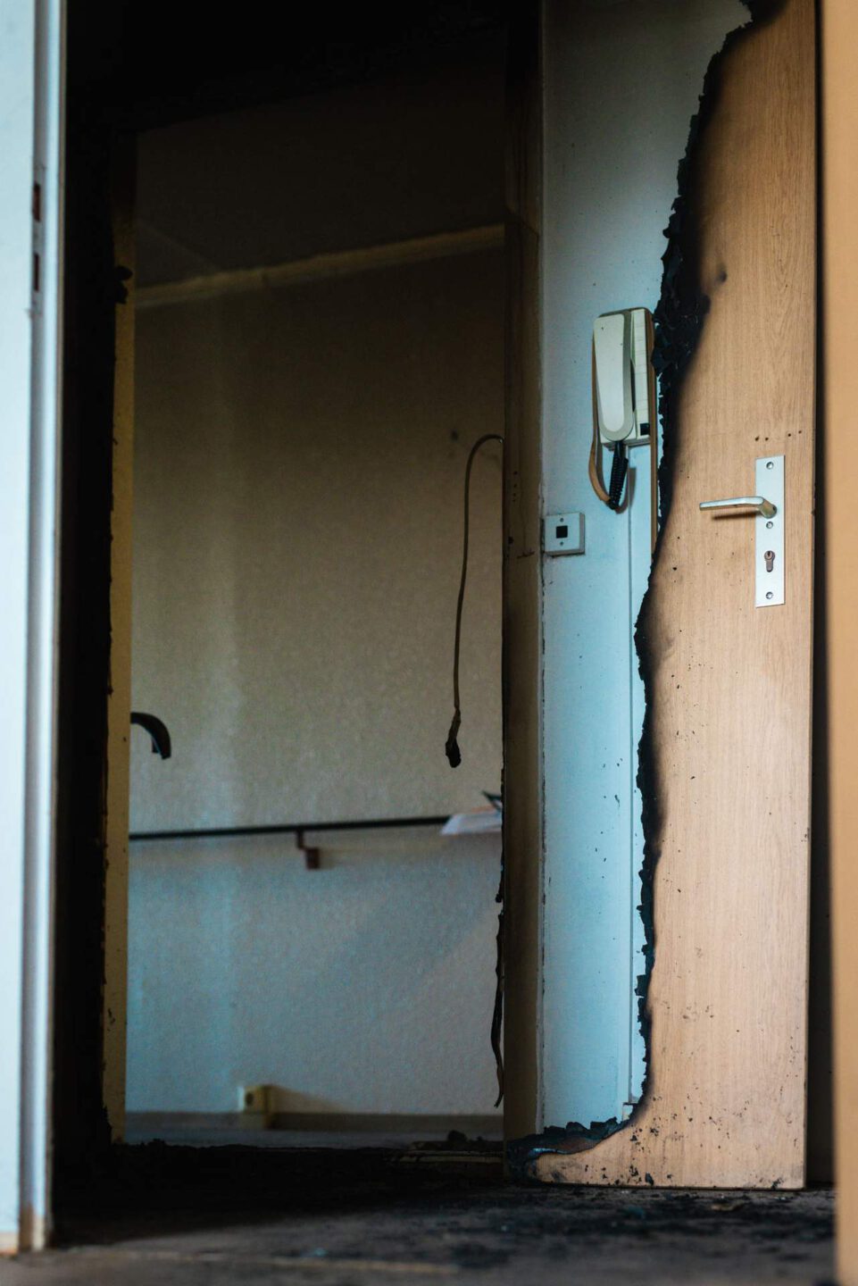 Blick durch eine offene, durch Brand beschädigte Tür in ein dunkles Zimmer. Der Türrand weist starke Rußschäden auf. Rechts ist eine Gegensprechanlage und ein Stecker an der Wand sichtbar.