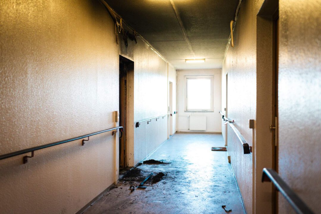 Langaufnahme eines langen Flurs in einem Wohn- oder Geschäftsgebäude, der durch einen Brand stark beschädigt wurde. Der Boden ist nass und teilweise mit Ruß bedeckt. Am Ende ist ein helles Fenster sichtbar.