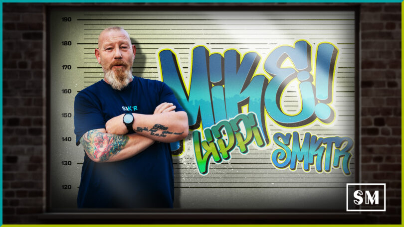 Mugshot-Stil Porträt von Mike, Mitarbeiter von SM Kattner, mit verschränkten Armen vor einer Messlatte und Graffiti-Schriftzug. Der Mann trägt ein dunkles T-Shirt mit "SMKR" Logo und hat Tätowierungen. Der Hintergrund ist eine Mauer.