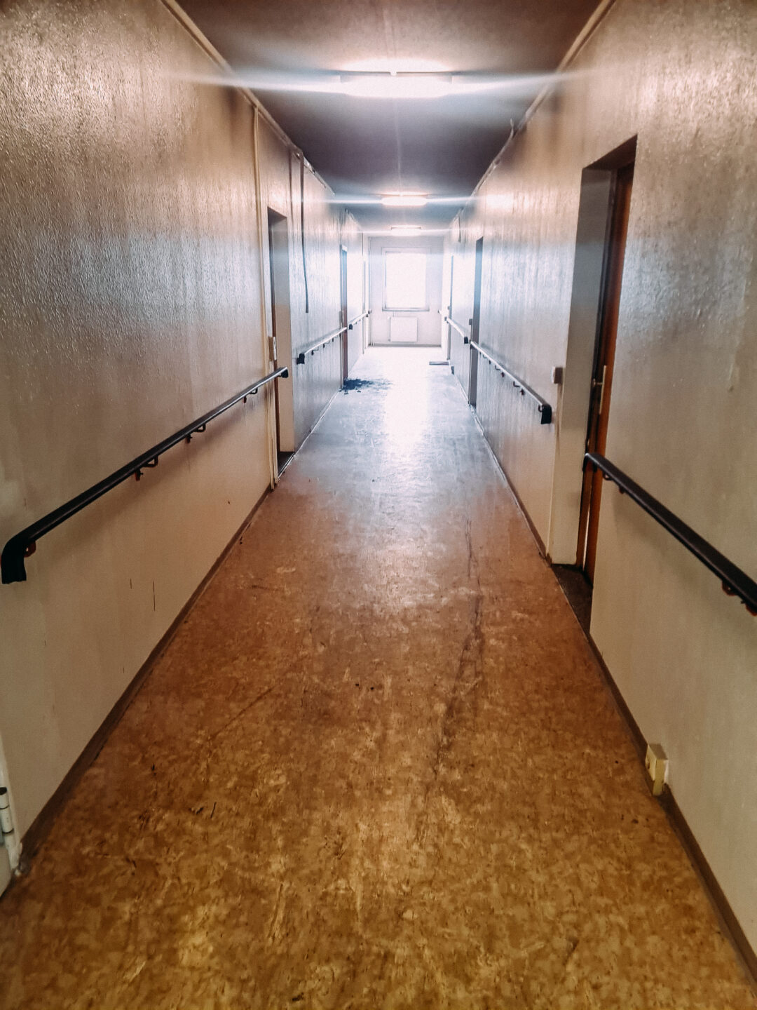 Länglicher, dunkler Flur eines Gebäudes, möglicherweise nach einem Brand oder mit Schimmelbefall. Die Wände sind hell und glänzend, der Boden ist braun/gelblich und schmutzig. Auf beiden Seiten des Flurs sind schwarze Handläufe montiert. Am Ende des Ganges scheint helles Licht durch ein Fenster, das den Schmutz auf dem Boden akzentuiert.