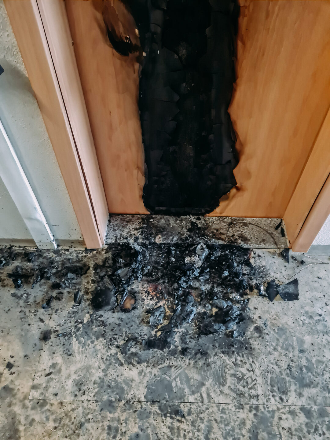 Detailansicht des Bodens direkt vor einer Tür, die von einer großen, vertikalen Brandspur gezeichnet ist. Auf dem Boden liegt eine dicke Schicht aus schwarzer Asche und verkohltem Material, die sich mit grauen und nassen Flecken des ursprünglichen Bodens vermischt.