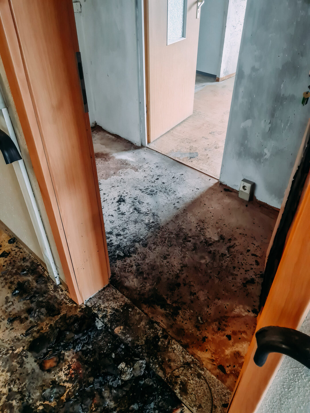 Nahaufnahme eines Türbereichs, der auf einen weiteren Raum führt. Der Boden ist mit schwarzen Brand- und Schmutzspuren übersät. Links und rechts begrenzen helle Holztürrahmen den Blick auf den dahinterliegenden, helleren Raum mit einer weiteren Tür.