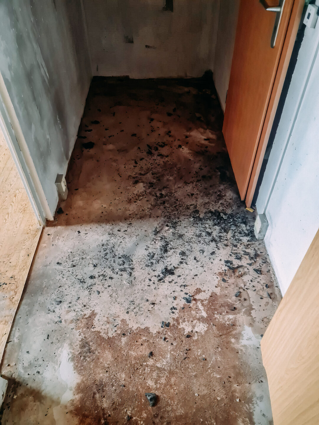 Blick nach unten auf einen kleinen, dunklen Flurbereich. Der Boden ist mit schwarzen Brandrückständen und Asche bedeckt, vermischt mit nassen, rötlich-braunen Flecken. Rechts ist der untere Teil einer hellbraunen Holztür zu sehen, links eine helle Wand.
