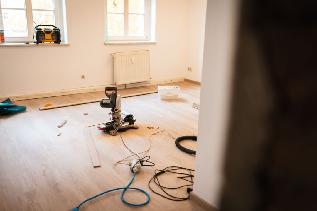 Eine Kappsäge steht auf dem hellen Holzboden in der Mitte eines leeren, weißen Raumes, der renoviert wird. Kabel liegen auf dem Boden. Im Hintergrund sind zwei Fenster, ein Radio und ein Heizkörper.