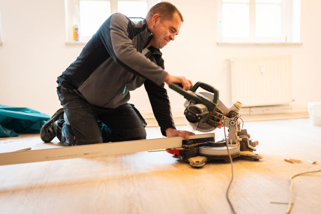 Ein Bauarbeiter kniet auf einem hellen Holzboden in einem Zimmer und benutzt eine Kappsäge, um eine lange, weiße Fußleiste zuzuschneiden. Im Hintergrund sind zwei Fenster und ein Heizkörper zu sehen.