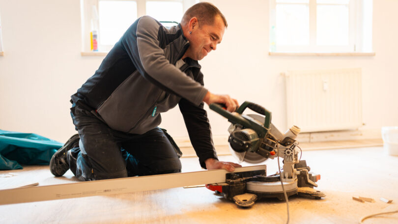 Ein Bauarbeiter kniet auf einem hellen Holzboden in einem Zimmer und benutzt eine Kappsäge, um eine lange, weiße Fußleiste zuzuschneiden. Im Hintergrund sind zwei Fenster und ein Heizkörper zu sehen.