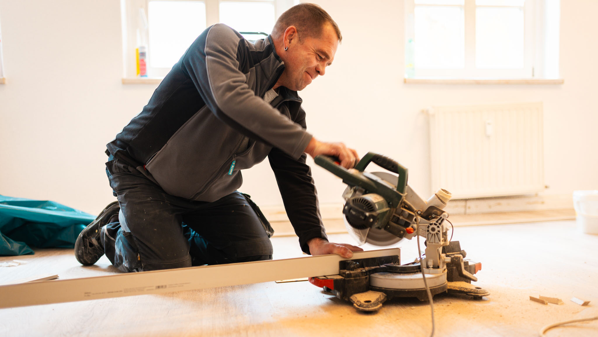 Ein Bauarbeiter kniet auf einem hellen Holzboden in einem Zimmer und benutzt eine Kappsäge, um eine lange, weiße Fußleiste zuzuschneiden. Im Hintergrund sind zwei Fenster und ein Heizkörper zu sehen.