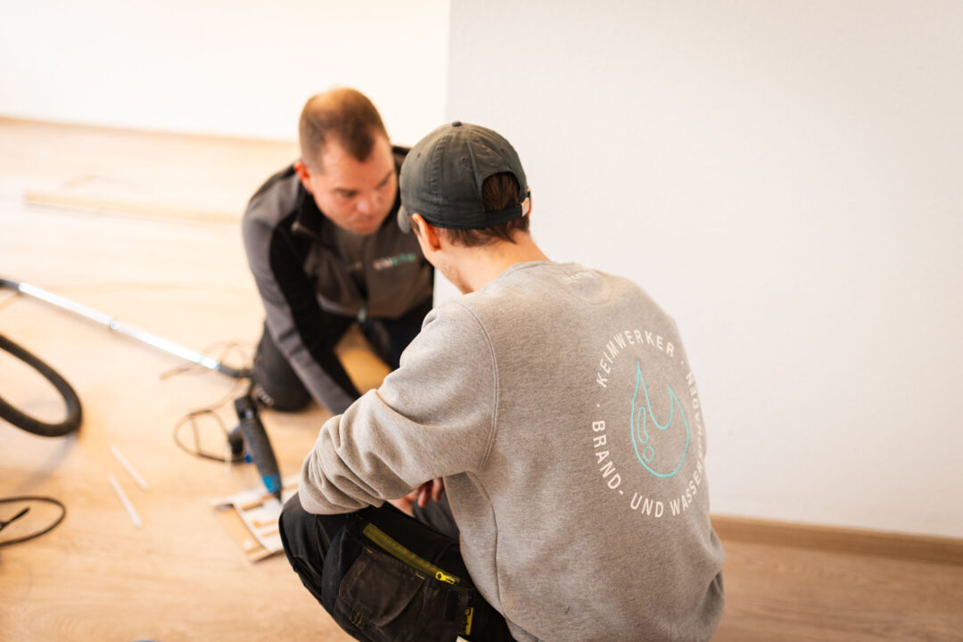 Zwei Bauarbeiter knien auf dem Boden in einem hellen Raum. Der Arbeiter im Vordergrund ist von hinten zu sehen und trägt einen grauen Pullover mit einem Logo auf dem Rücken, das eine stilisierte Wassertropfenform und den Text "HEIMWERKER BRAND- UND WASSERSCHADEN" zeigt. Der andere Arbeiter bückt sich neben ihm.