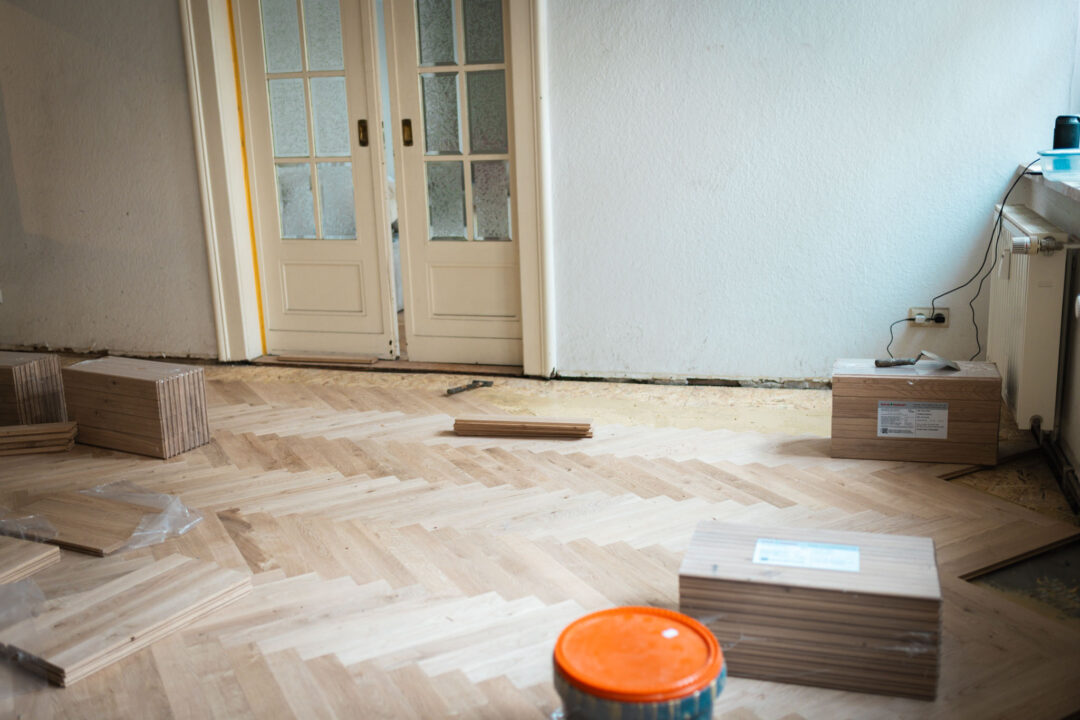 Ein Raum, in dem bereits ein Teil des neuen Fischgrät-Parketts verlegt wurde. Auf dem Boden stehen gestapelte Holzkisten mit weiteren Dielen und ein großer, oranger Eimer. Im Hintergrund ist eine doppelte, weiße Glastür zu sehen.