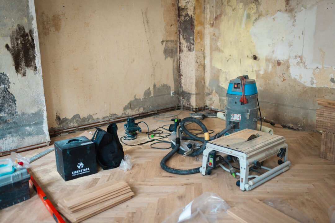 Eine Ecke eines Raumes in Renovierung. Die Wände sind alt, mit abgeblättertem Putz und Verfärbungen. Auf dem Fischgrät-Parkett liegen Baustellenwerkzeuge, darunter ein großer, blauer Industriestaubsauger, eine Tischsäge, Kabel, eine schwarze Werkzeugtasche und Stapel neuer Parkettdielen.