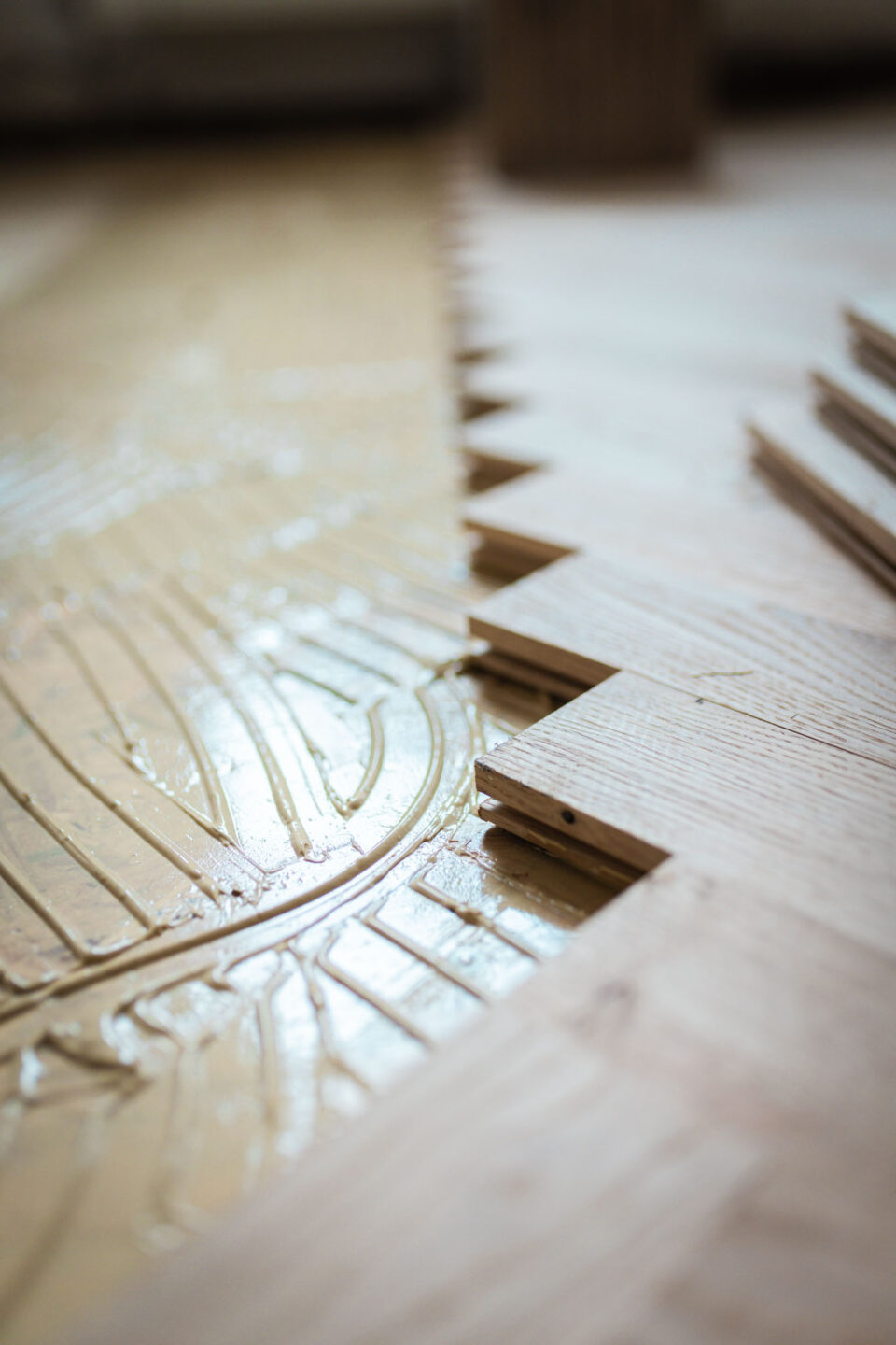 Nahaufnahme des Bodens, auf dem Parkettdielen im Fischgrätmuster verlegt werden. Die frisch aufgetragene, dicke Schicht Klebstoff mit Rillen ist deutlich zu sehen, während neue Holzstücke in die Klebefläche eingesetzt werden.