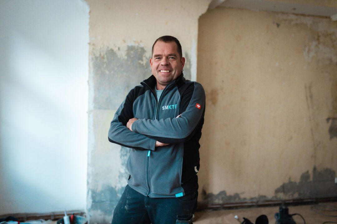 Ein lächelnder Bauarbeiter, Sven, in grauer und schwarzer Arbeitskleidung (Jacke mit der Aufschrift "SMKTE") steht mit verschränkten Armen in einem renovierungsbedürftigen Raum. Die Wand im Hintergrund ist teilweise hell verputzt und teilweise rau und gelblich.