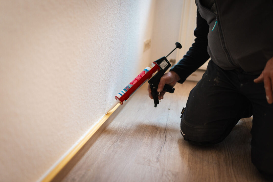 Nahaufnahme eines Bauarbeiters, der kniet und eine Kartuschenpresse benutzt, um eine weiße Silikonnaht zwischen dem hellen Holzboden und der weißen Wand anzubringen.
