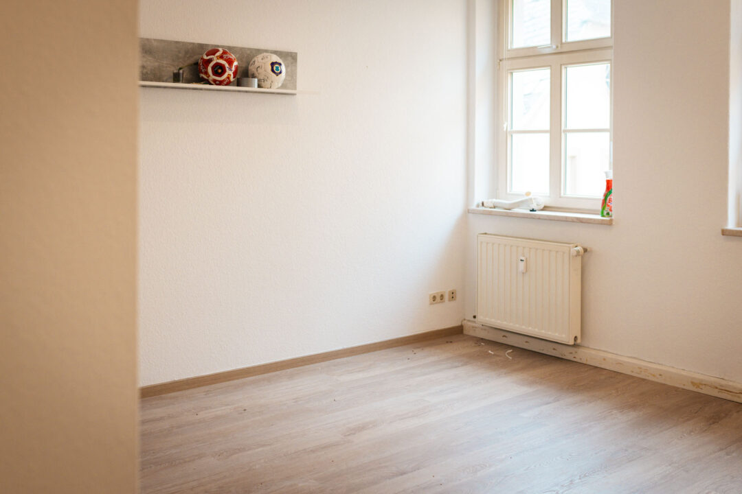Ein fertig renovierter, leerer, weißer Raum mit hellem Holzboden. Es ist ein Fenster mit einem Heizkörper darunter zu sehen. An der Wand hängt ein kleines Regal mit zwei dekorativen Fußbällen darauf. Der Bodenrand ist mit Fußleisten versehen.