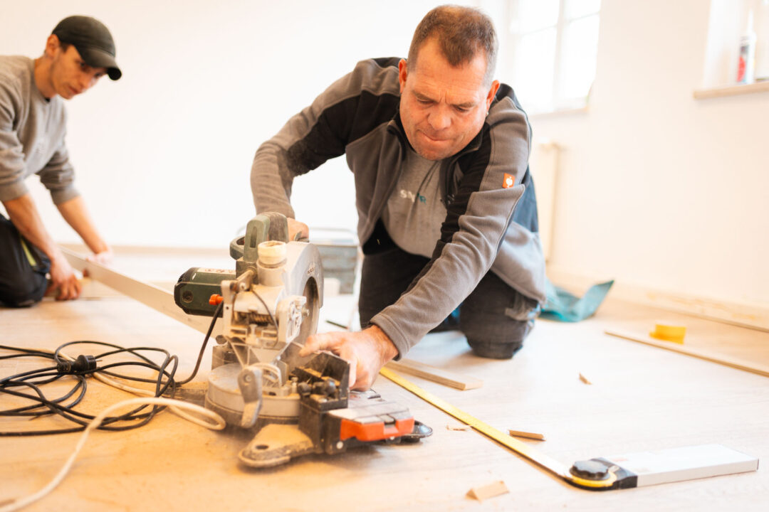 Zwei Handwerker bei der Bodenrenovierung: Fokus auf Mitarbeiter im Vordergrund, der eine elektrische Gehrungssäge bedient, während ein Kollege im Hintergrund assistiert.
