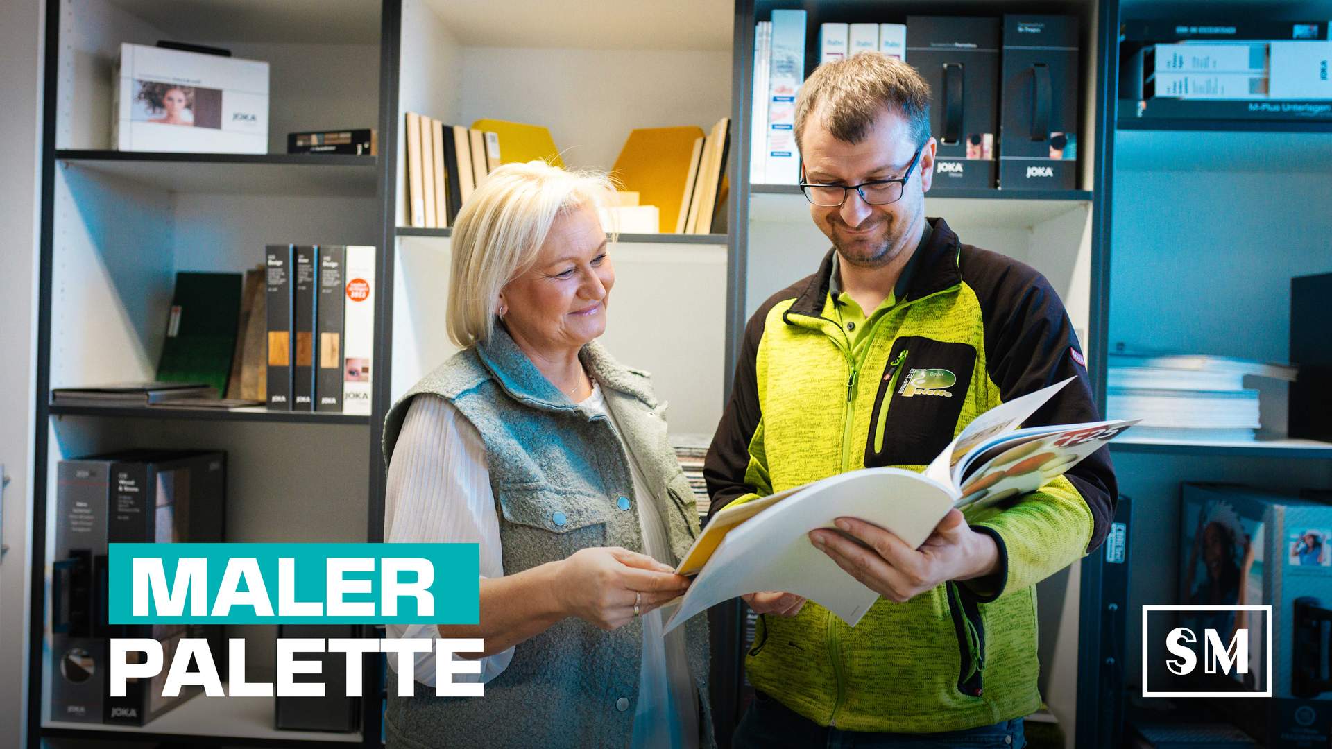 Zwei Mitarbeiter der Malerpalette schauen sich vor einem Regal voller Ordner einen Musterkatalog ihrer Firma an.