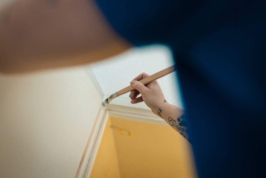 Eine tätowierte Hand streicht mit einem Pinsel präzise die Kante zwischen einer gelben Wand und einer weißen Decke.