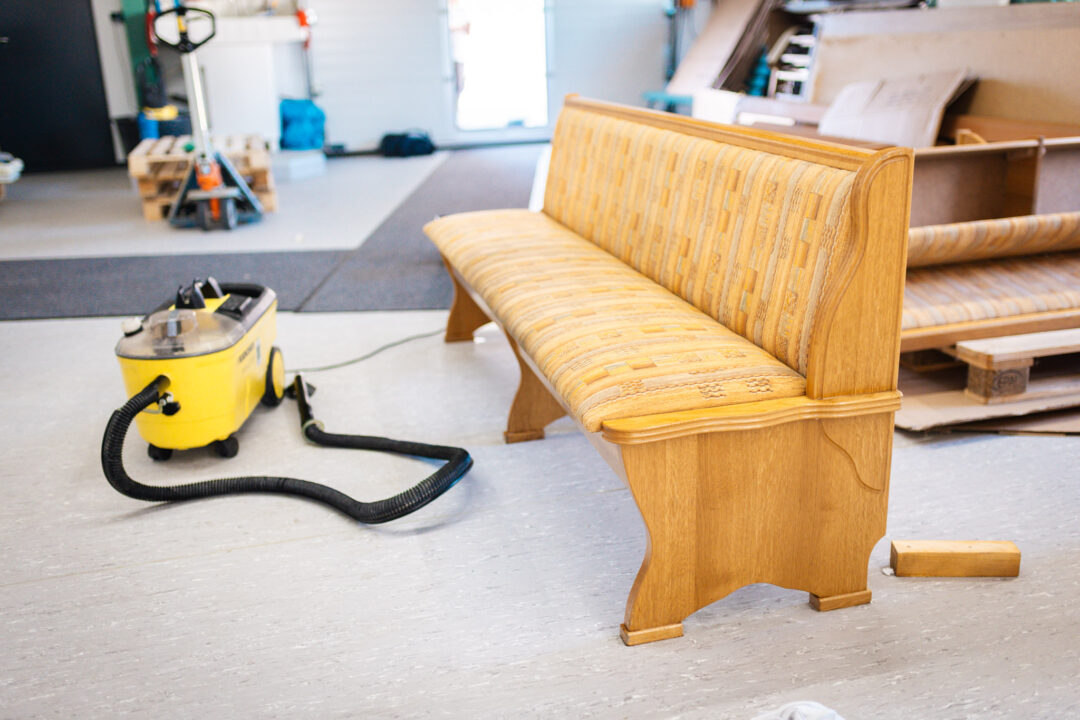 Ein heller Werkstattraum, in dem eine hölzerne Eckbank mit gemusterten Polstern für die Sanierung bereitsteht. Im Vordergrund befindet sich ein gelber Sprühextraktionssauger (Profi-Waschsauger) mit ausgelegtem Schlauch, der für die Tiefenreinigung der Textilien nach dem Kellerbrand genutzt wird. Im Hintergrund ist weiteres Werkstattequipment wie ein Hubwagen zu erkennen
