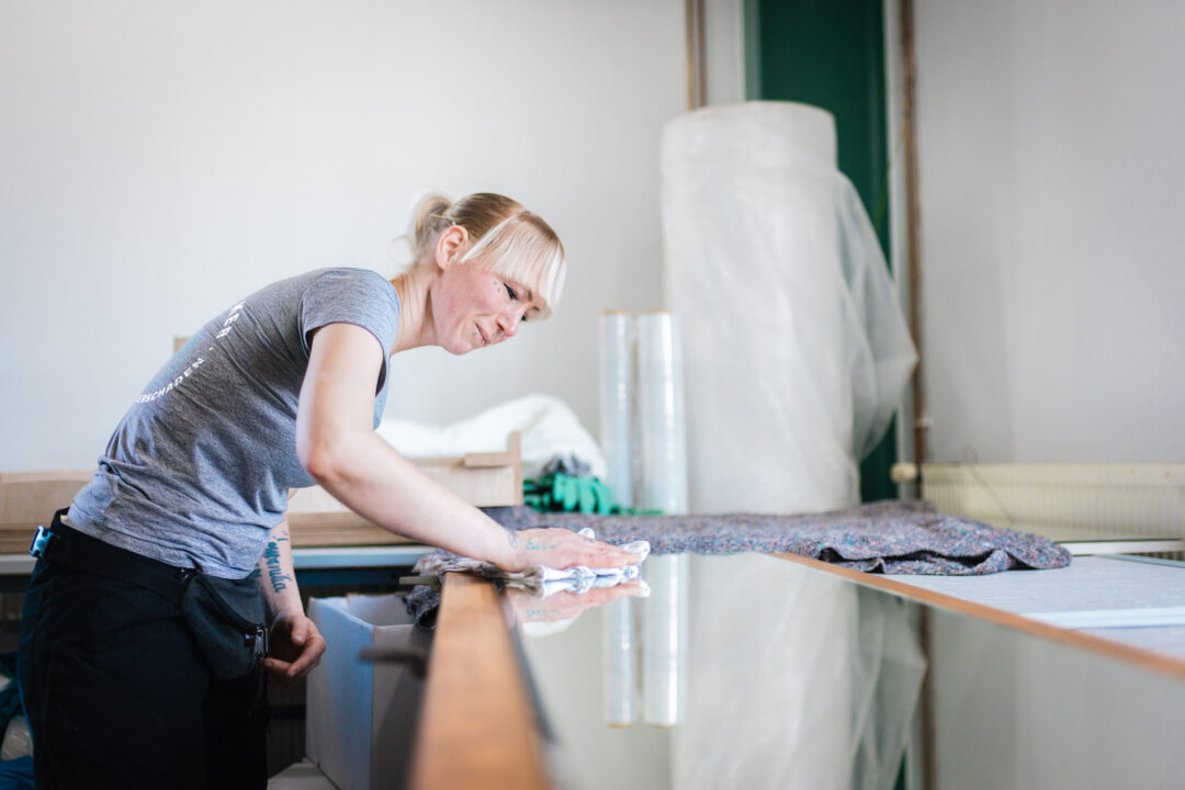 Eine Mitarbeiterin von SM Kattner wischt sorgfältig mit einem Reinigungstuch über die Kante eines Möbelstücks, um Rußpartikel und Brandrückstände zu entfernen. Sie trägt Arbeitskleidung mit dem Firmenlogo; im Hintergrund ist Verpackungsmaterial wie Luftpolsterfolie für den geschützten Rücktransport der gesäuberten Möbel zu sehen.