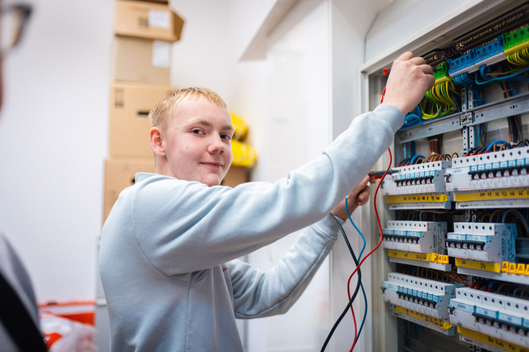 Ein junger Auszubildender der Elektro Beck GmbH prüft lächelnd mit Messleitungen die Verdrahtung in einem geöffneten Schaltschrank.