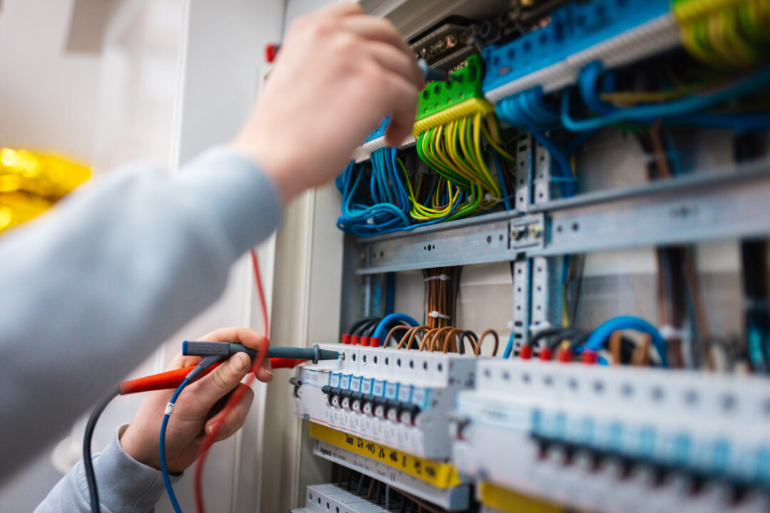 Detailansicht der Elektroinstallation: Ein Elektriker nutzt Prüfspitzen an den Klemmen eines Sicherungsblocks zur Fehlersuche und Qualitätskontrolle.