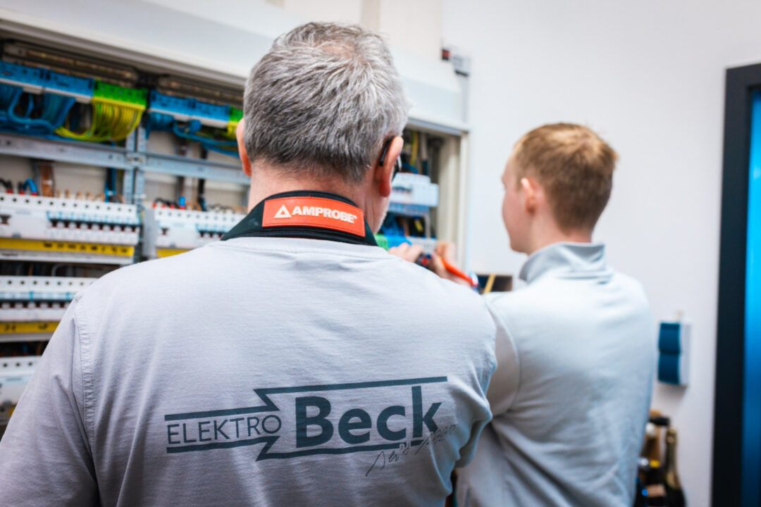 Rückansicht eines erfahrenen Elektromeisters im grauen Firmen-T-Shirt der Elektro Beck GmbH bei der gemeinsamen Arbeit mit einem Lehrling am Schaltkasten.