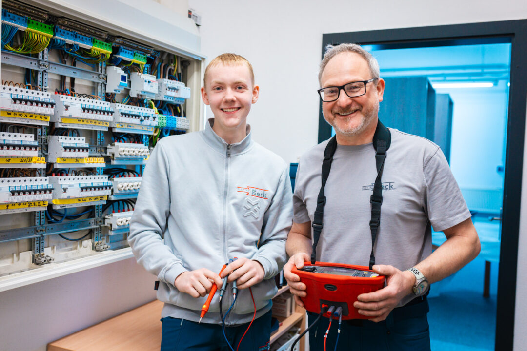 Ein freundlicher Lehrling und ein erfahrener Elektriker der Elektro Beck GmbH stehen lächelnd mit Messwerkzeugen vor einem fertig installierten Schaltschrank im Hauptquartier.