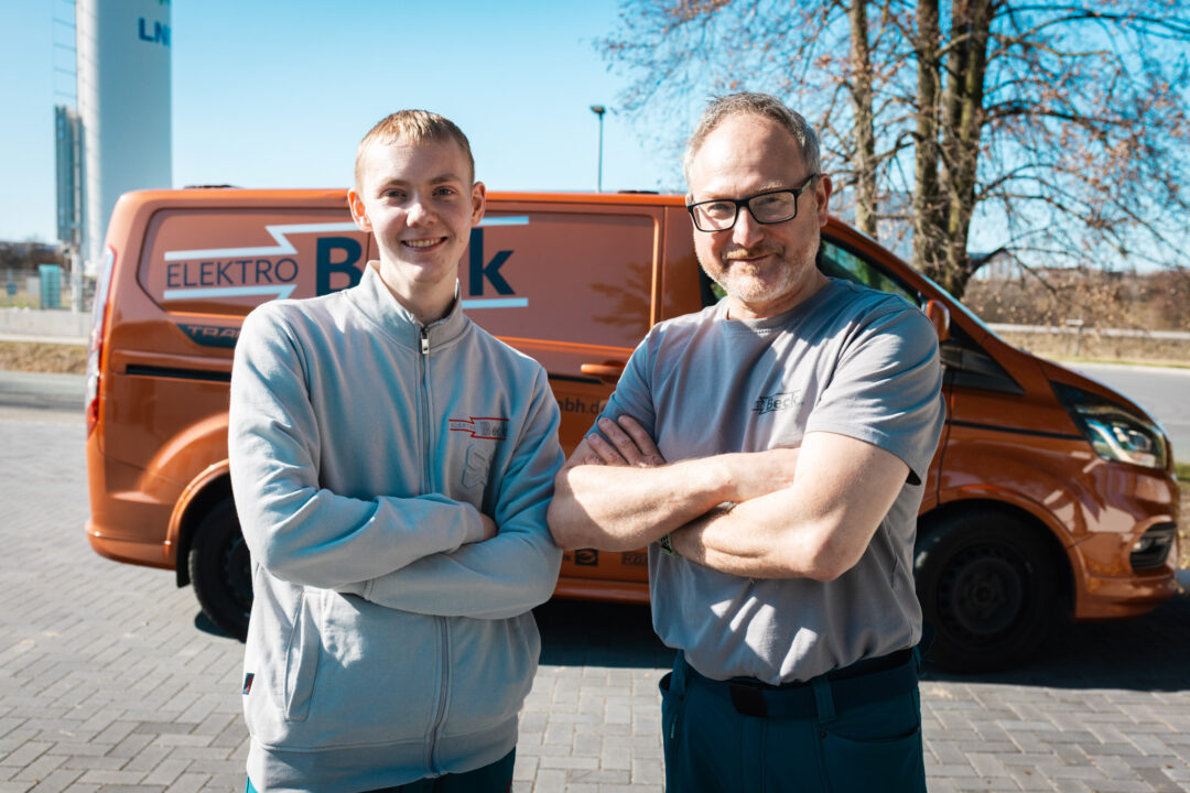 Zwei Generationen Elektro-Handwerk: Zwei Mitarbeiter der Elektro Beck GmbH posieren mit verschränkten Armen vor dem orangefarbenen Firmen-Transporter.