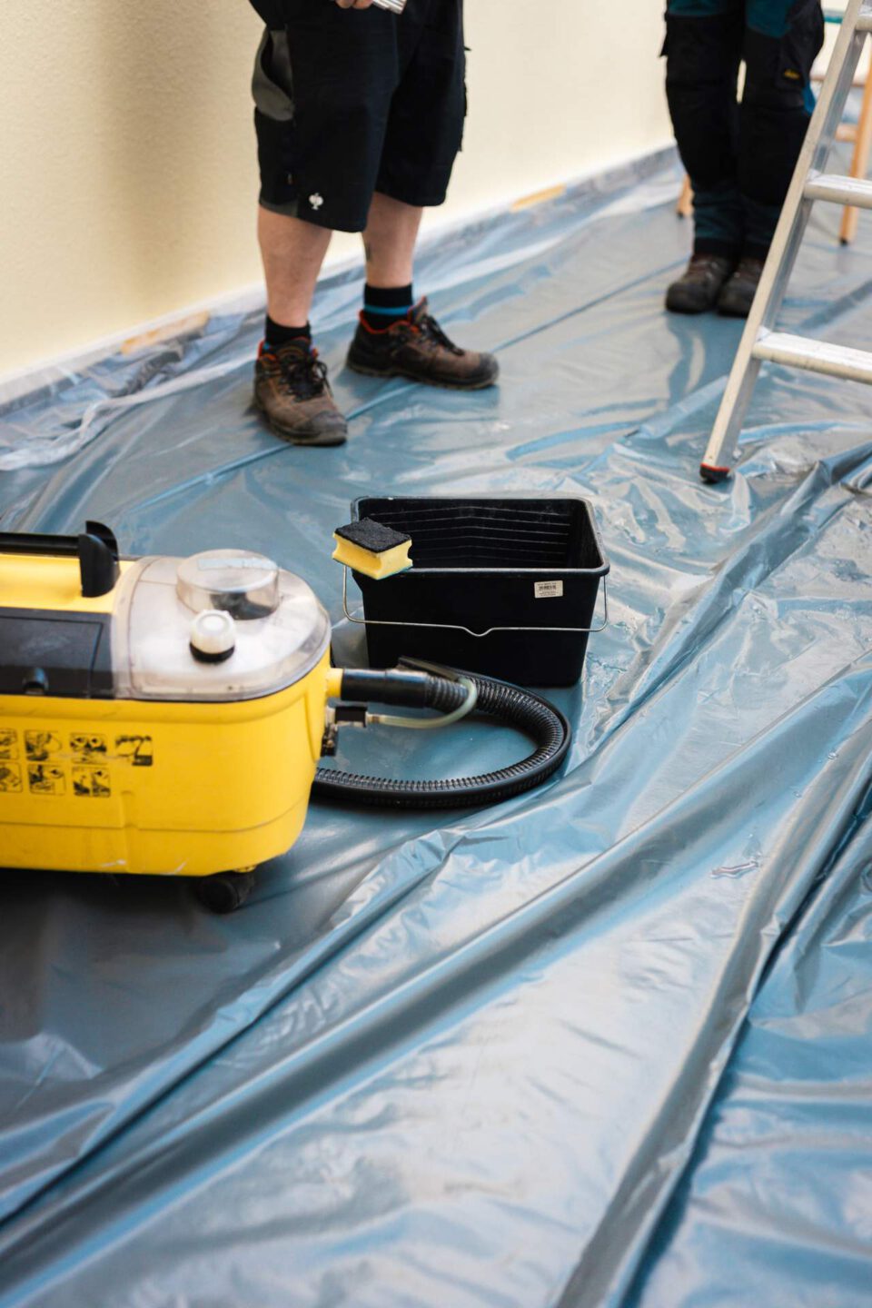 Reinigungsequipment für die Rußbeseitigung, bestehend aus einem gelben Nassauger, einem schwarzen Eimer und einem Schwamm, steht auf einer ausgelegten Schutzfolie auf dem Boden.