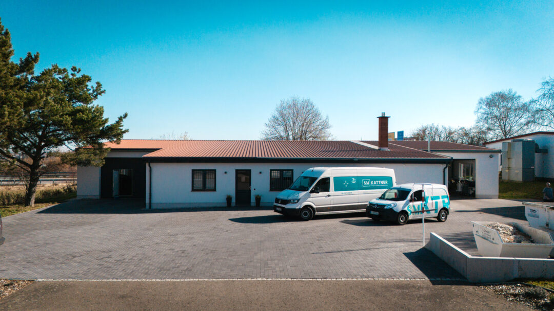 Weitwinkel-Luftaufnahme des Betriebsgebäudes der SM Kattner GmbH mit gepflastertem Hof und zwei Firmenfahrzeugen.