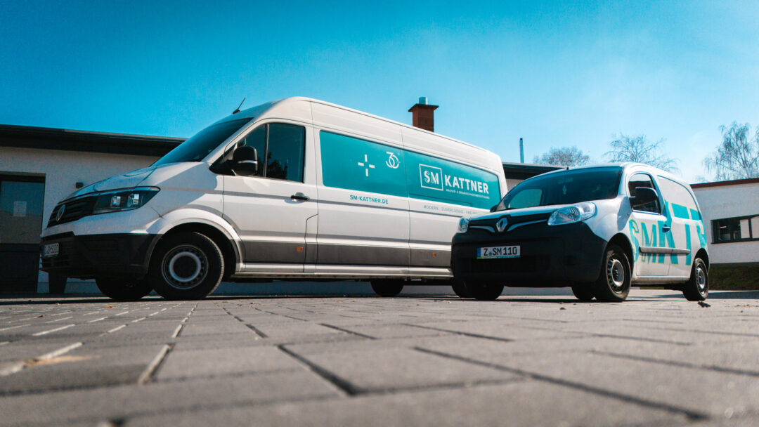 Fotografie aus der Froschperspektive (Bodennähe): Ein weißer VW-Transporter und ein kleinerer Renault-Firmenwagen der SM Kattner GmbH stehen nebeneinander auf einem gepflasterten Hof. Beide Fahrzeuge tragen die cyan/petrolfarbene Firmenbeschriftung 'SM Kattner - Wasser- & Brandschadensanierung'. Im Hintergrund ist ein blauer, wolkenloser Himmel zu sehen.