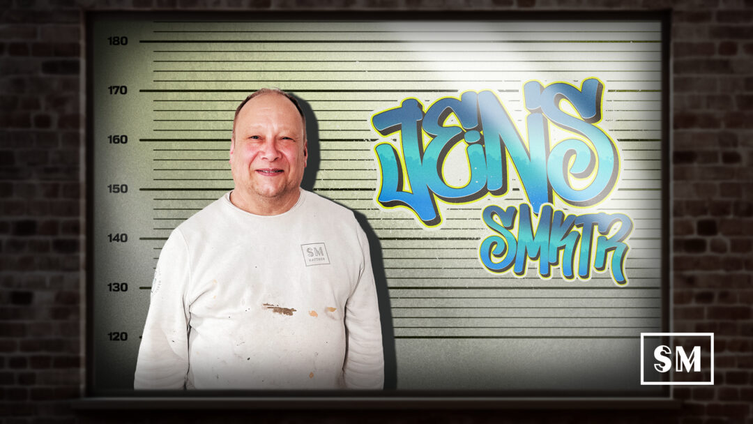 Mitarbeiter Jens lächelt vor einer Polizeifoto-Hintergrundwand mit Graffiti-Schriftzug Jens SM KTR.