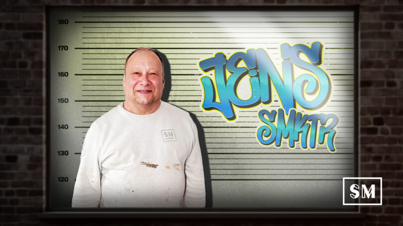 Mitarbeiter Jens lächelt vor einer Polizeifoto-Hintergrundwand mit Graffiti-Schriftzug Jens SM KTR.